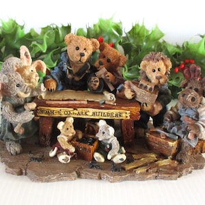 May include: A detailed figurine depicting animal characters, including bears, an elephant, and a rabbit, gathered around a wooden table. The table has the text "NOAH & CO. ARK BUILDERS". The figurine is set on a brown base with green foliage in the background.