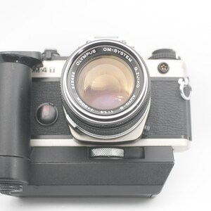 May include: A silver and black Olympus OM-System camera with a Zuiko Auto-S 1:1.8 50mm lens attached. The camera has a black hand grip and a black flash unit.