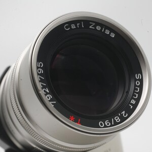 May include: Close-up of a silver Carl Zeiss Sonnar 2.8/90 camera lens. The lens has black lettering that reads "Carl Zeiss Sonnar 2.8/90" and "1927795". The lens is angled towards the viewer.