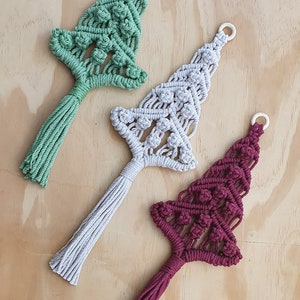 May include: Three macrame wall hangings shaped like Christmas trees in sage green, light gray, and deep burgundy. Each tree features intricate knotted patterns, a small wooden ring at the top for hanging, and a long, straight fringe at the base.