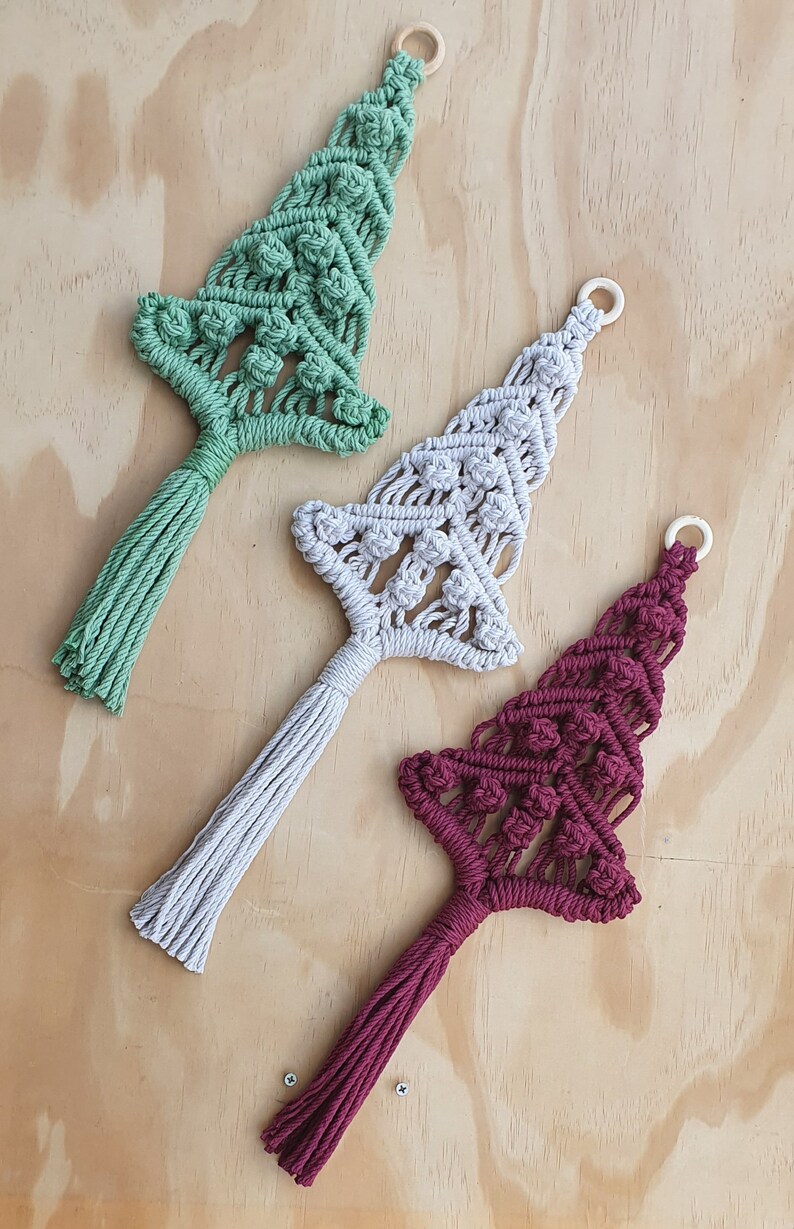 May include: Three macrame wall hangings shaped like Christmas trees in sage green, light gray, and deep burgundy. Each tree features intricate knotted patterns, a small wooden ring at the top for hanging, and a long, straight fringe at the base.