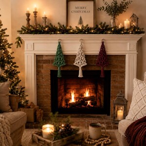 May include: Three macrame Christmas tree ornaments in green, cream, and burgundy hanging from a white fireplace mantel. Each ornament features intricate knotted cord patterns and a bottom tassel, displayed above a lit fireplace with flames.