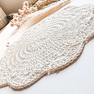 Puede incluir: Una alfombra redonda de crochet blanca con un borde festoneado. La alfombra está hecha de hilo grueso y tiene una superficie texturizada.