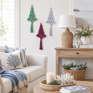 May include: Three macrame wall hangings shaped like Christmas trees in sage green, deep magenta, and light gray, each featuring intricate knotted patterns and a long tassel at the bottom, suspended from small wooden rings on a white wall.