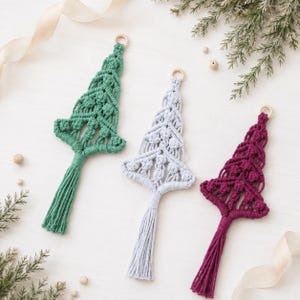 May include: Three macrame ornaments shaped like Christmas trees in green, light gray, and deep magenta. Each ornament features intricate knotted patterns, a wooden ring at the top for hanging, and a long, straight tassel at the bottom base.