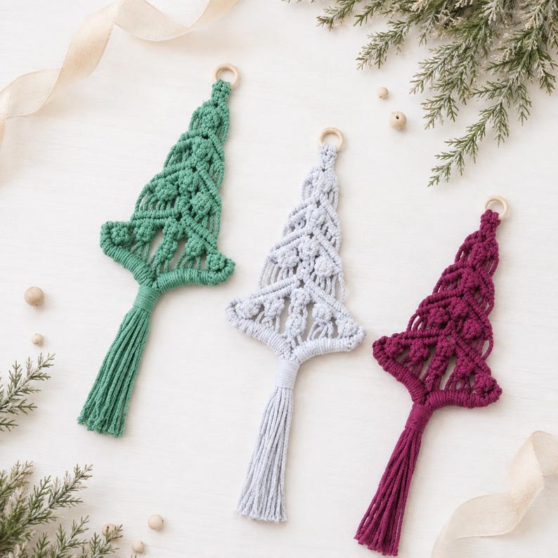 May include: Three macrame ornaments shaped like Christmas trees in green, light gray, and deep magenta. Each ornament features intricate knotted patterns, a wooden ring at the top for hanging, and a long, straight tassel at the bottom base.