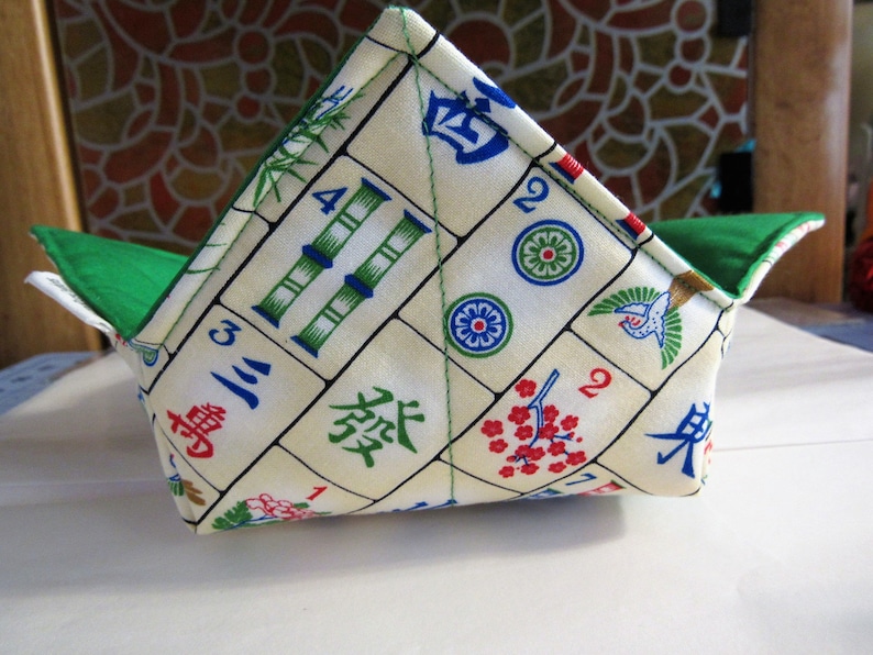 May include: A green and white fabric bowl with a Mahjong tile pattern. The bowl is folded into a triangular shape.