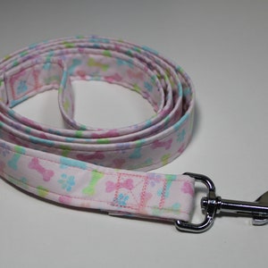 May include: A rolled-up dog leash with a light pink background and a repeating pattern of colorful bones and paw prints. The leash has a silver metal clasp.