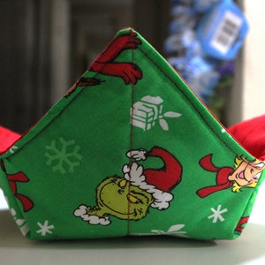May include: A green and red fabric bowl cozy featuring a Christmas-themed design. The exterior is green with images of a cartoon character in a Santa hat and snowflakes. The interior is red. The cozy is designed to hold a bowl.