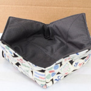May include: A square, fabric bowl cozy with a black interior and a patterned exterior. The exterior features a white background with colorful, Asian-inspired symbols. The cozy is designed to hold a bowl and keep food warm.