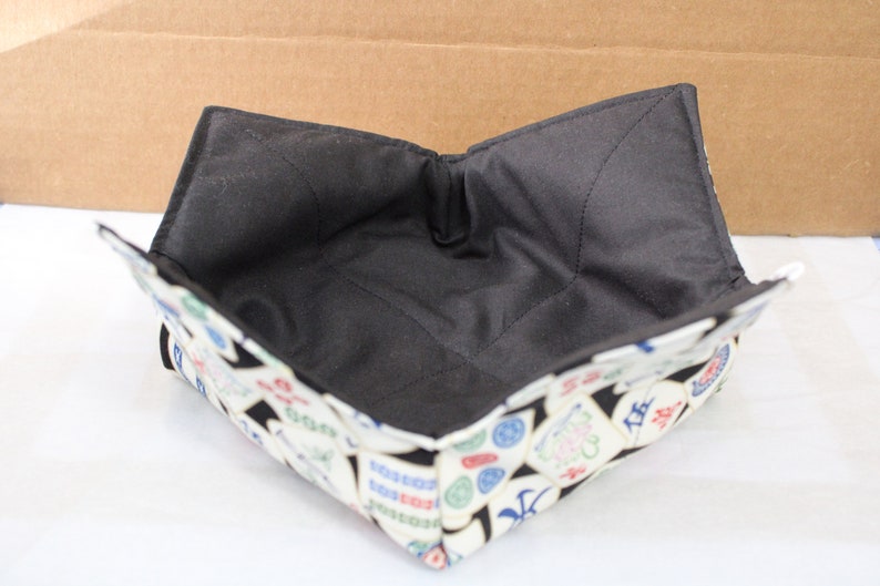 May include: A square, fabric bowl cozy with a black interior and a patterned exterior. The exterior features a white background with colorful, Asian-inspired symbols. The cozy is designed to hold a bowl and keep food warm.
