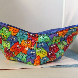 May include: A blue fabric bowl with a colorful cat print. The cats are in various poses and colors, including red, orange, yellow, green, blue, and purple.