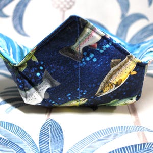 May include: A square fabric bowl with a blue fish pattern. The bowl has a dark blue interior with images of fish and bubbles. The edges are folded up to create a bowl shape. The background is a blue and white floral pattern.