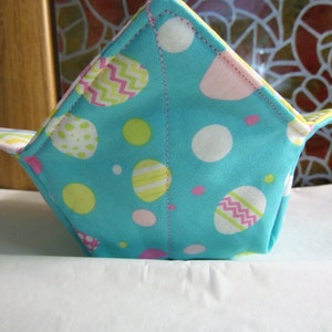 May include: A turquoise fabric bowl cozy with an Easter egg pattern in white, pink, and yellow. The cozy is square-shaped with folded corners and a contrasting patterned interior. The stitching is visible along the edges.