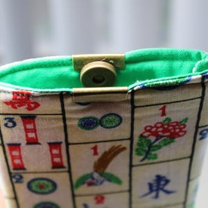May include: A fabric pouch with a green interior and a brass clasp. The exterior features a pattern of Mahjong tiles with red, green, and blue accents. A small keychain is attached to the side.