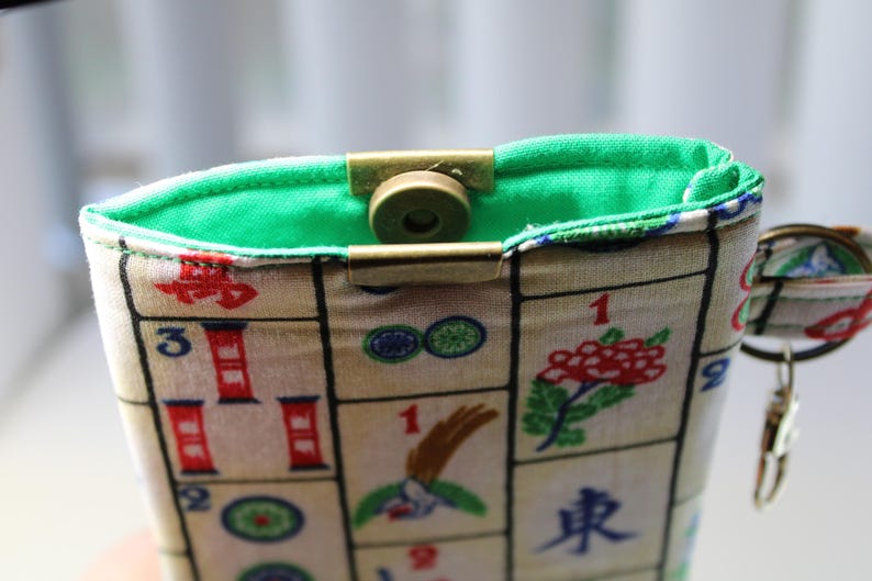 May include: A fabric pouch with a green interior and a brass clasp. The exterior features a pattern of Mahjong tiles with red, green, and blue accents. A small keychain is attached to the side.