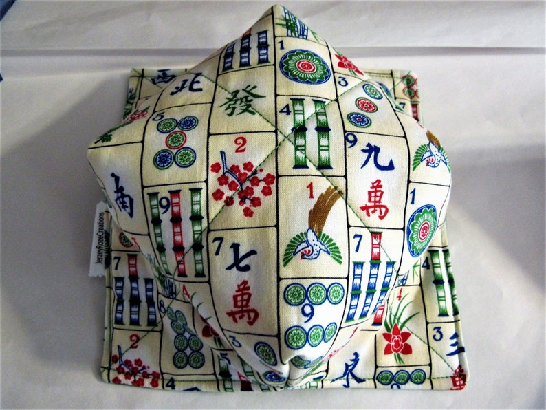May include: A white fabric square with a pattern of mahjong tiles, numbers, and Chinese characters. The fabric is quilted and has a 3D effect. The fabric is folded into a cube shape.