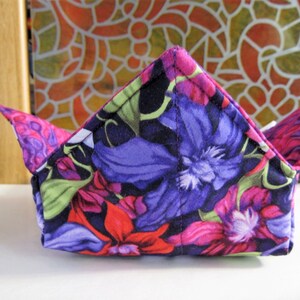 May include: A folded fabric bowl with a floral print in shades of purple, pink, and green. The bowl is made of cotton fabric and has a triangular shape.