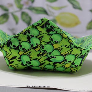 May include: A green and yellow fabric bowl with a repeating pattern of cartoon alien faces. The bowl is square-shaped with four upturned corners, creating a unique, open design. The background is a blurred image of lemons and leaves.