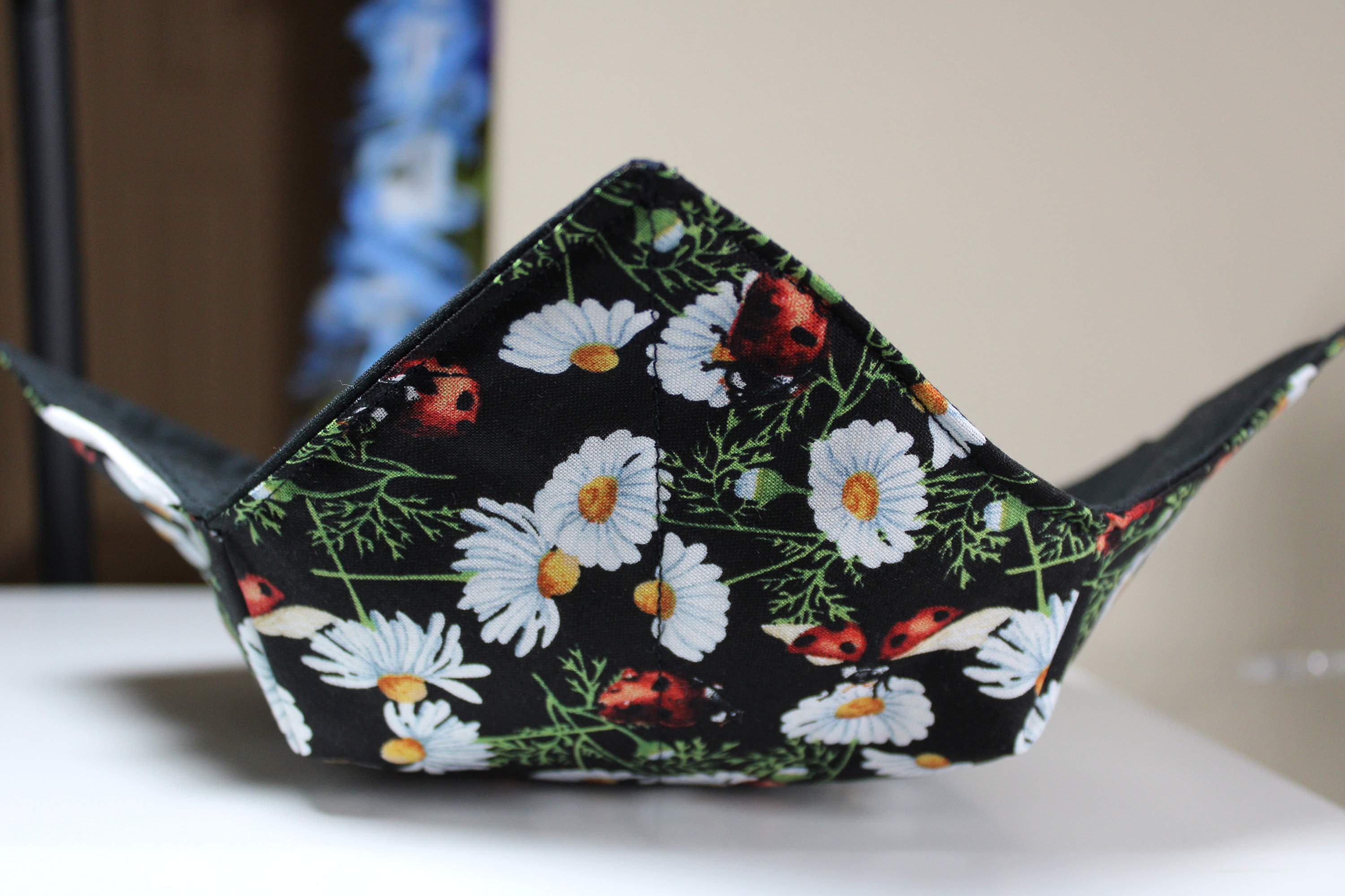 May include: A black fabric bowl with a floral pattern featuring white daisies and red ladybugs. The bowl is folded and appears to be empty.