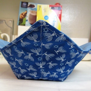 May include: A blue fabric bowl with a white menorah and Star of David pattern. The interior is a solid light blue. The bowl is open and square-shaped, designed to hold food items.