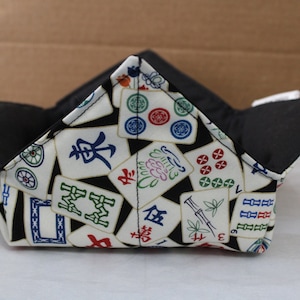 May include: A fabric bowl with a black interior and a white exterior patterned with colorful Mahjong tiles. The bowl is folded into a square shape with four upward-pointing corners.