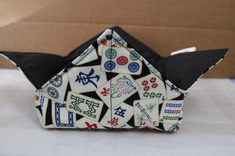 May include: A fabric bowl with a black interior and a white exterior patterned with colorful Mahjong tiles. The bowl is folded into a square shape with four upward-pointing corners.