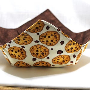 Peut inclure: Un bol en tissu brun et blanc avec un motif de biscuits aux pépites de chocolat. Le bol est plié et a une doublure brune.