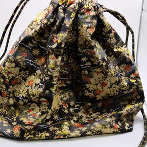 May include: A black drawstring bag featuring a dense, intricate pattern of gold, red, and white floral and leaf motifs. The fabric has a subtle sheen, and the bag is cinched at the top with two black and gold braided cords that serve as handles.