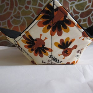 May include: A fabric bowl cozy with a turkey and floral pattern in brown, orange, and yellow on a cream background. The interior is black, and the text "GOBBLE Til You Wobble" is printed on the side.