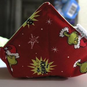 May include: A red fabric bowl with a green Grinch design. The bowl has a green and white lining and the text "The Grinch" is printed on the front.