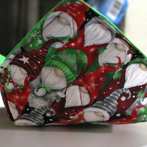 May include: A fabric bowl cozy with a festive gnome pattern in red, green, and gray. The interior is bright green. The cozy is designed to hold a bowl of hot food, protecting hands from heat. A holiday-themed kitchen accessory.