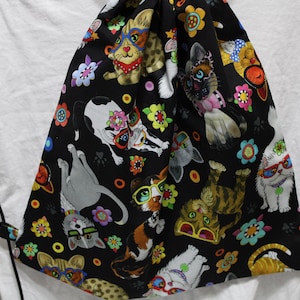Peut inclure: Sac à cordon noir avec un motif de chats colorés. Les chats portent des lunettes de soleil et sont entourés de fleurs. Le sac est en tissu et possède des cordons noirs.