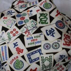 May include: A pair of fabric bowl cozies featuring a repeating pattern of colorful Mahjong tiles on a black background. The tiles have red, green, blue, and gold accents. The cozies are designed to hold bowls.
