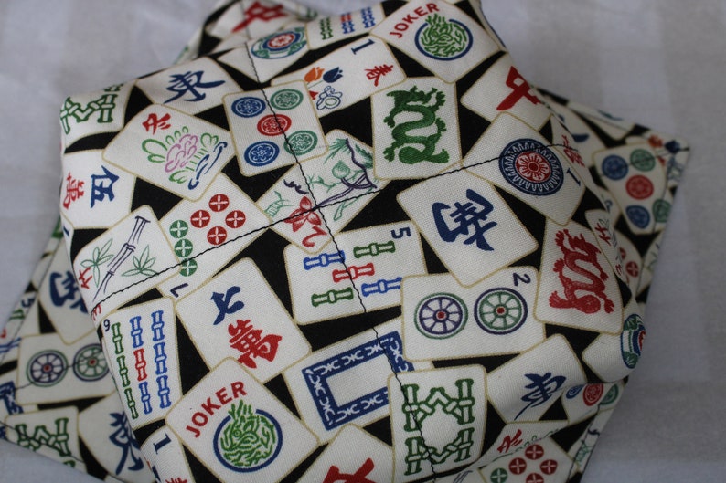 May include: A pair of fabric bowl cozies featuring a repeating pattern of colorful Mahjong tiles on a black background. The tiles have red, green, blue, and gold accents. The cozies are designed to hold bowls.