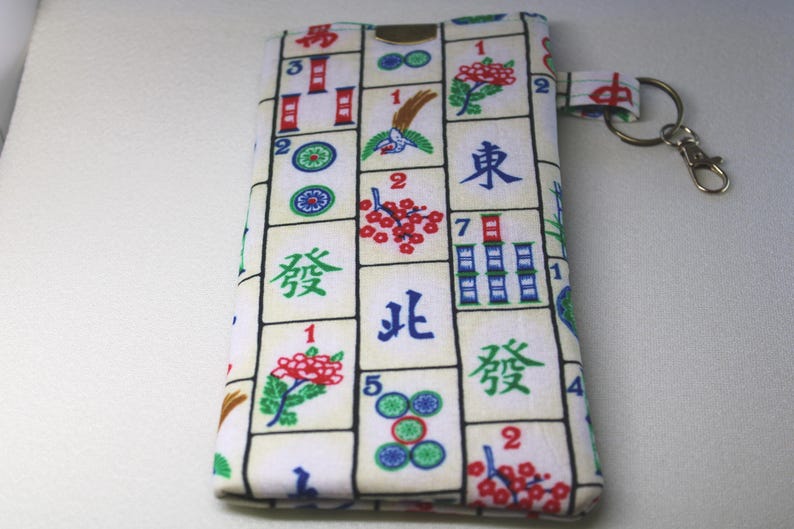 May include: A rectangular fabric pouch with a Mahjong tile pattern in red, green, blue, and black. The pouch has a small loop with a metal clasp and key ring attached. The background is a plain white surface.
