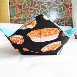 May include: A black fabric bowl with a blue lining and a pattern of pies and spoons. The bowl is folded and sitting on a white surface.