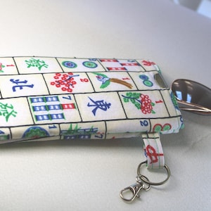 May include: A rectangular fabric pouch with a Mahjong tile pattern in white, green, red, and blue. A small, silver scoop is next to the pouch. The pouch has a small strap with a silver key ring.