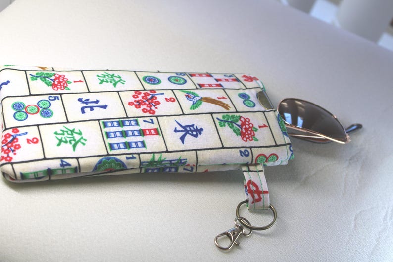 May include: A rectangular fabric pouch with a Mahjong tile pattern in white, green, red, and blue. A small, silver scoop is next to the pouch. The pouch has a small strap with a silver key ring.
