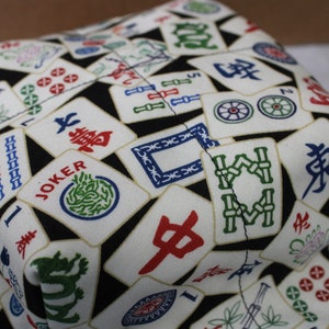 May include: A fabric item with a black background covered in white tiles featuring colorful Chinese characters and symbols. The tiles include red, green, and blue designs, with the word "JOKER" visible on one tile. The item appears to be a container.