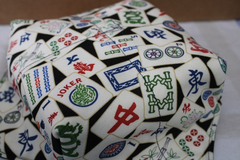 May include: A fabric item with a black background covered in white tiles featuring colorful Chinese characters and symbols. The tiles include red, green, and blue designs, with the word "JOKER" visible on one tile. The item appears to be a container.