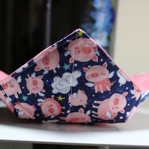 May include: A pink and blue fabric bowl with a pattern of flying pigs on a dark blue background. The bowl is folded and has a pink lining.