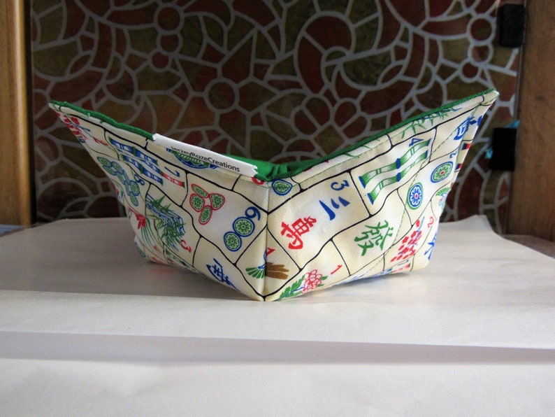 May include: A green and white fabric bowl with a Mahjong tile pattern. The bowl is folded and sitting on a white surface. The text "Pizza Creations" is visible on the bowl.
