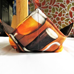 May include: A square bowl cozy with a beer-themed print. The fabric features images of beer glasses and mugs in shades of brown, orange, and white. The cozy has dark brown corners and edges, and is designed to hold a bowl of food.