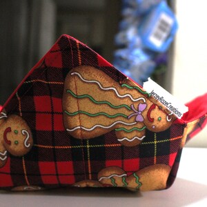 May include: A fabric bowl cozy with a red and black plaid pattern and gingerbread man designs. The interior is solid red. The cozy is designed to hold a bowl and keep food warm. The text "JerzeyRozeCreations" is visible.