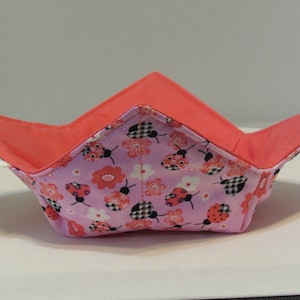 May include: A pink and coral fabric bowl with a ladybug and flower pattern. The bowl is folded flat and is made of a soft, quilted fabric.