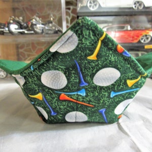 May include: A green fabric bowl with a golf theme. The bowl features a pattern of white golf balls and colorful tees on a green background. The interior and trim are solid green.
