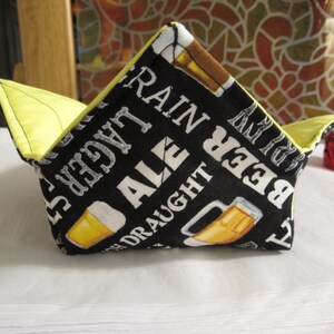 May include: A black fabric bowl with a yellow lining. The bowl has a beer theme with the words "Beer", "Ale", "Draught", "Lager", and "Rain" printed on it. There are also images of beer mugs printed on the bowl.