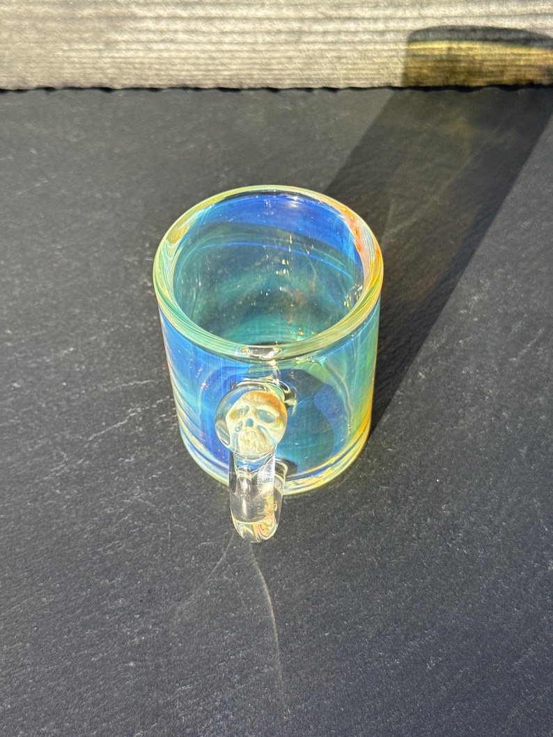 May include: A small glass mug featuring an iridescent, multi-colored finish that shifts between blue, yellow, and orange hues. The handle is made of clear glass and is molded into the shape of a small human skull at the top attachment point.