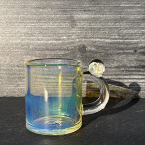 May include: A small, cylindrical glass mug featuring an iridescent finish that shifts between blue and yellow hues. The mug has a clear, curved handle with a small, spherical glass bead accent attached to the top of the handle connection point.